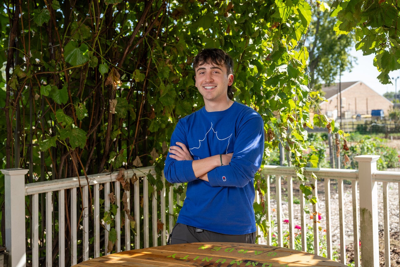 "A young White man with short brown hair wearing a blue long-sleeve shirt with a white sycamore leaf design stands smiling with his arms crossed under a leafy pergola in a garden area."