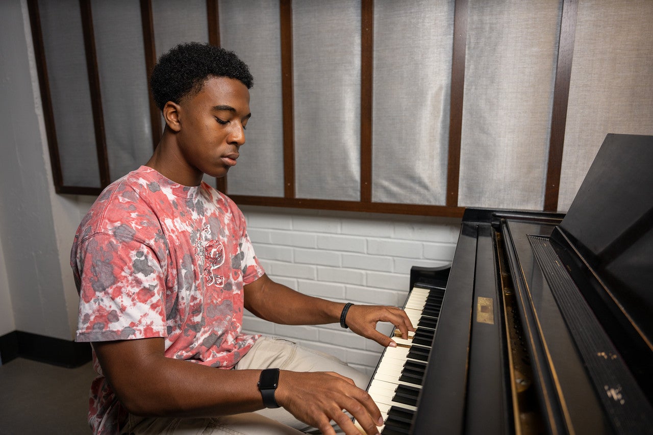 A young Black man wearing a red and white tie-dye T-shirt and a smartwatch plays an upright piano in a music room with white brick walls and vertical acoustic panels.