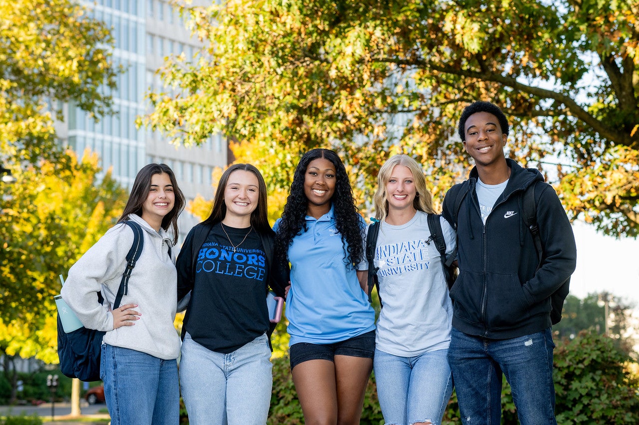 Five young adults of diverse racial backgrounds stand together outdoors in front of trees and a modern building, smiling and casually dressed with backpacks and college-themed T-shirts