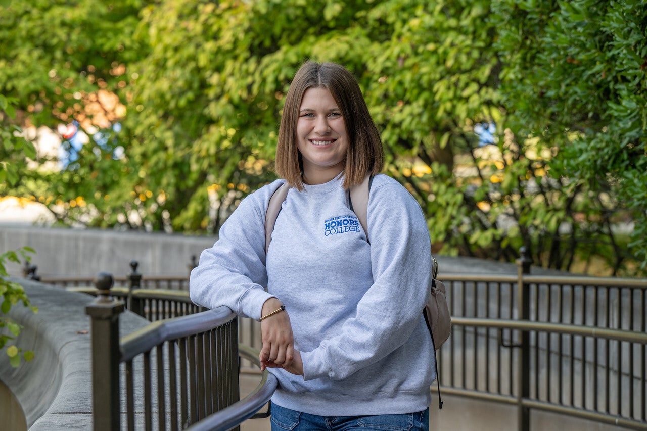 A young White woman with straight shoulder-length brown hair, wearing a light gray sweatshirt with “Indiana State University Honors College” written in blue on the front, blue jeans, and a beige backpack stands outdoors, smiling while leaning on a curved metal railing. Green leafy trees fill the background.