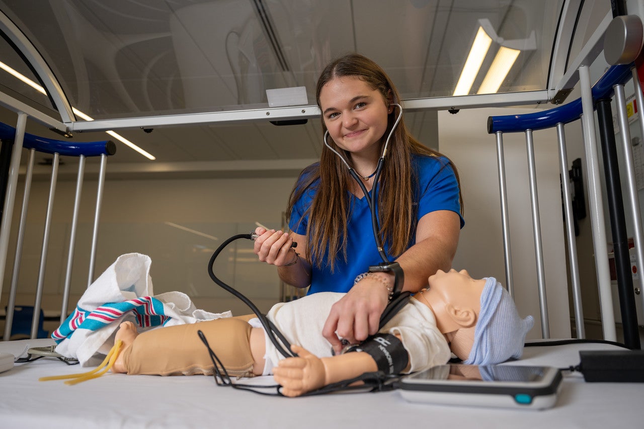A young white woman with long, straight brown hair, wearing a blue Indiana State University Nursing uniform, uses a stethoscope on a baby mannequin in a medical simulation lab.