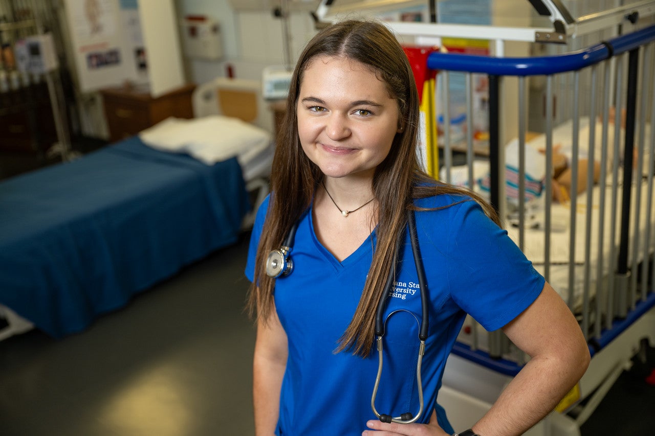 A young white woman with long, straight brown hair wearing a blue Indiana State University Nursing uniform and a stethoscope around her neck smiles while standing in a medical simulation lab with a hospital bed and equipment in the background.