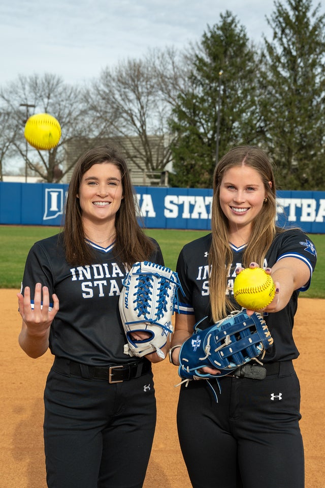 Two young White women wearing black Indiana State University softball uniforms with blue and white trim stand on a softball field smiling. Each holds a blue or white softball glove, and one tosses a yellow softball in the air while the other presents one forward.