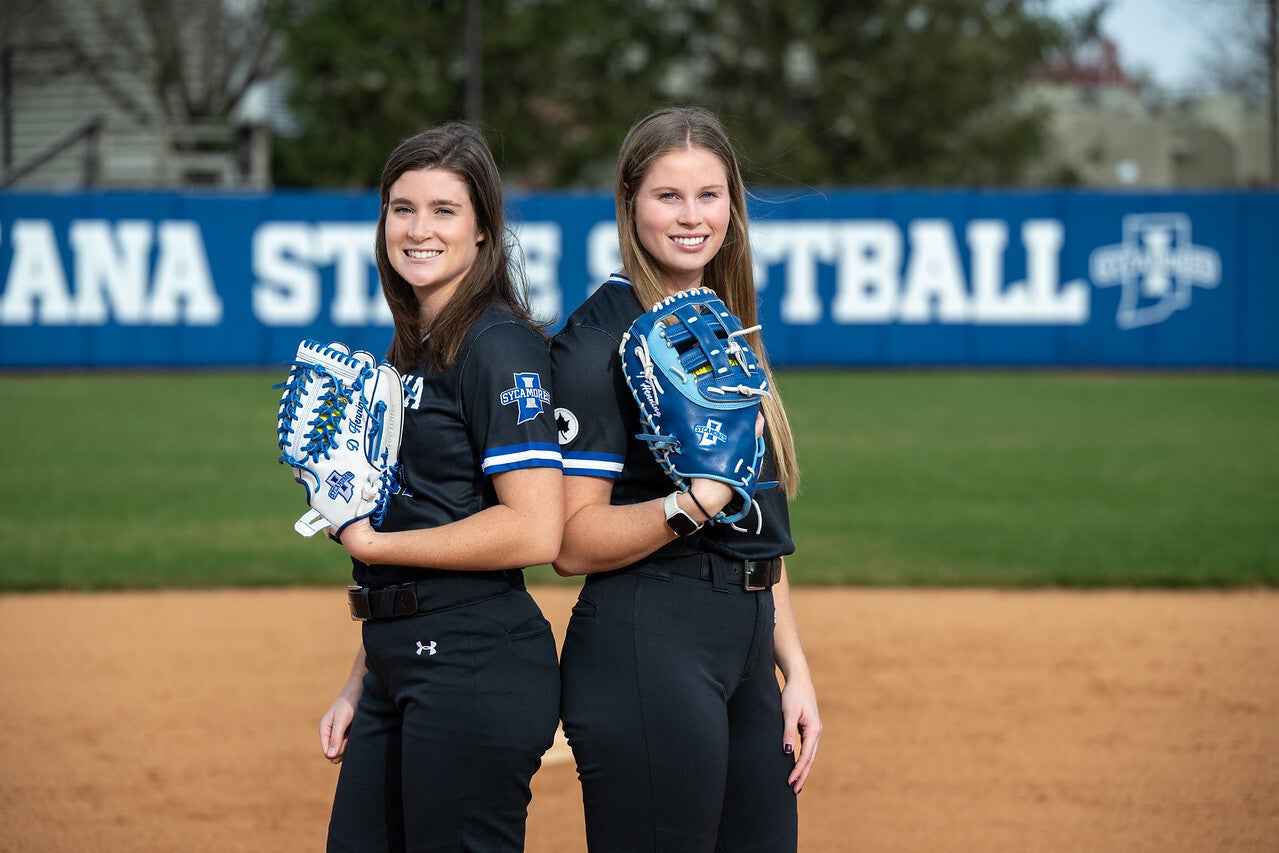 Two young White women stand back-to-back on a softball field, wearing black Indiana State University softball uniforms with blue accents. Each holds a blue or white softball glove, standing in front of a blue banner that reads “Sycamores Softball.”