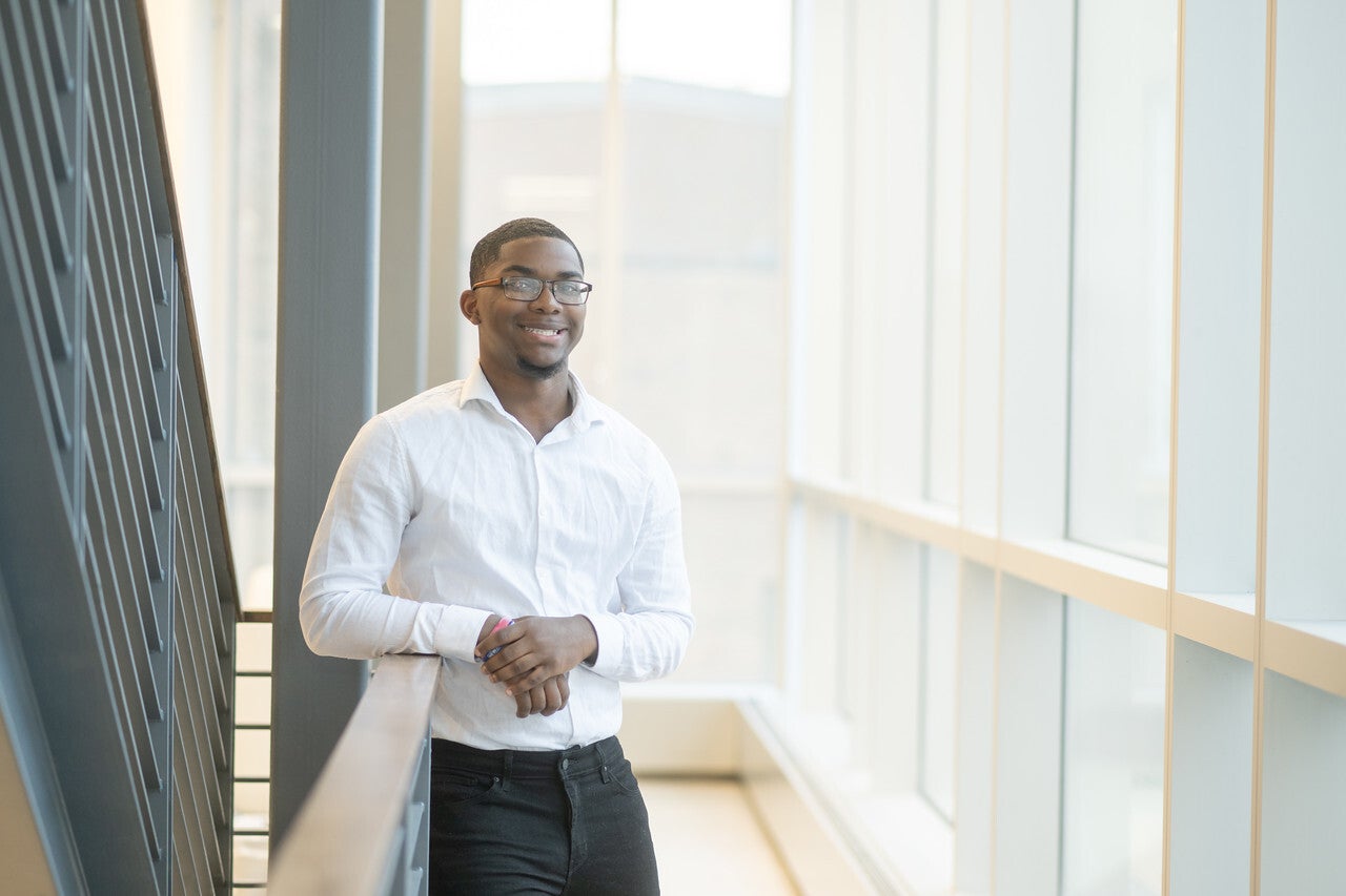 A smiling Black man wearing glasses, a white collared shirt, and dark pants leans on a railing in a bright hallway with large windows.