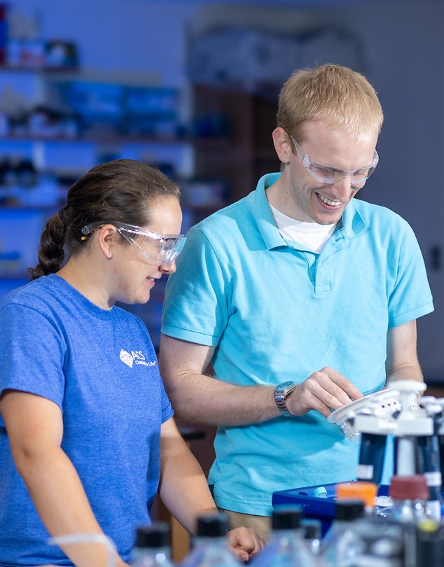 A white woman in a blue “ASC Chemistry Dept.” t-shirt and a white man in a light blue polo, both wearing safety goggles, smile while working with lab equipment. The table holds bottles and a white apparatus, with shelves of lab supplies in the background.