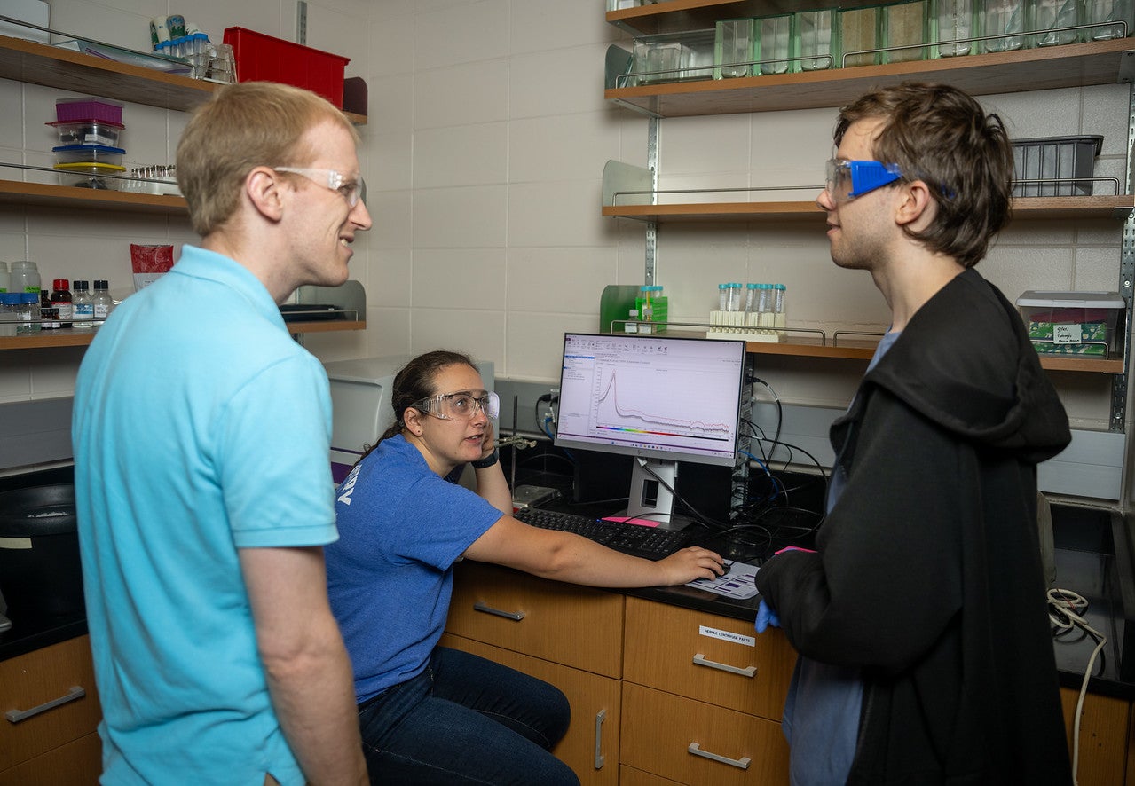 A young white woman sits at a computer displaying a multicolored graph, while two young white men stand nearby in conversation. All three wear safety goggles in a lab filled with bottles, test tubes, and containers.