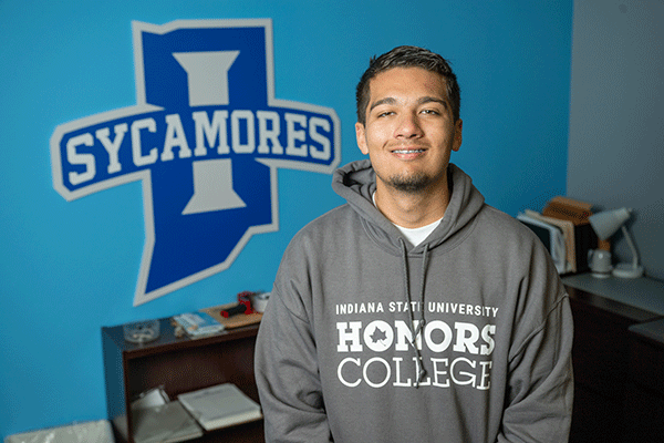 A smiling male student with short, dark hair is standing indoors in front of a blue wall featuring a large Indiana State University Sycamores logo. He is wearing a gray hoodie with the text 'INDIANA STATE UNIVERSITY HONORS COLLEGE.' A desk with books and papers is visible in the background.”