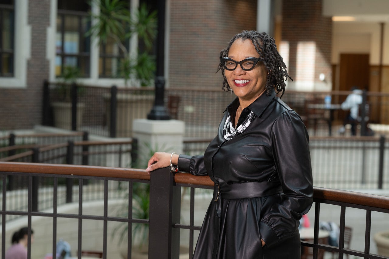 A smiling Black woman with short twists and black glasses stands indoors, leaning on a railing. She wears a long black dress with a belt and a black-and-white beaded necklace. A brick building and plants appear in the background.
