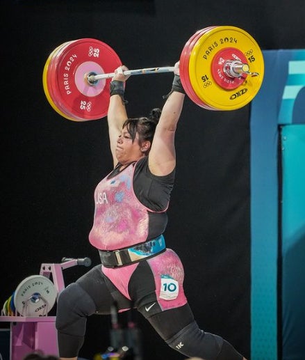 A female weightlifter in a pink and black “USA” uniform holds a loaded barbell overhead during a clean and jerk. Red plates read “PARIS 2024.” She wears a lifting belt and a “10” sticker on her left thigh