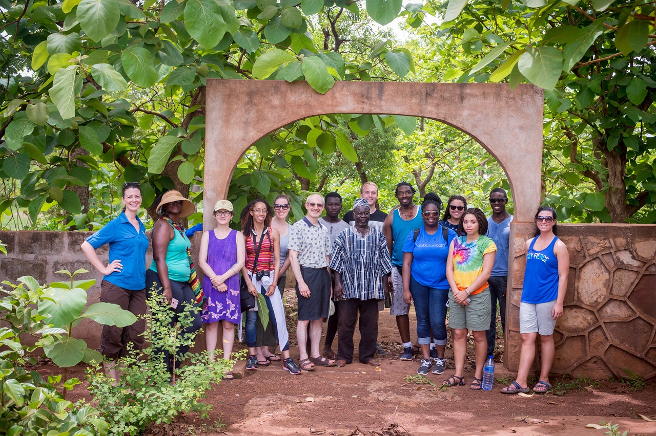 A diverse group of men and women stand on a dirt path under a stone arch surrounded by lush green trees. The group includes Black and White individuals dressed in casual summer clothing, such as T-shirts, tank tops, shorts, and hats