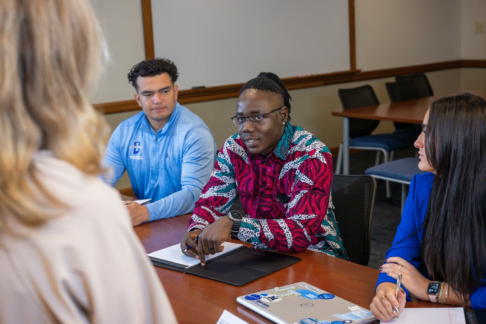 A Black male student with braided hair and glasses, wearing a red patterned shirt, speaks during a classroom discussion with a Latino man in a light blue shirt and a White woman with straight dark hair in a blue blouse, while another White woman with blonde wavy hair stands facing them.