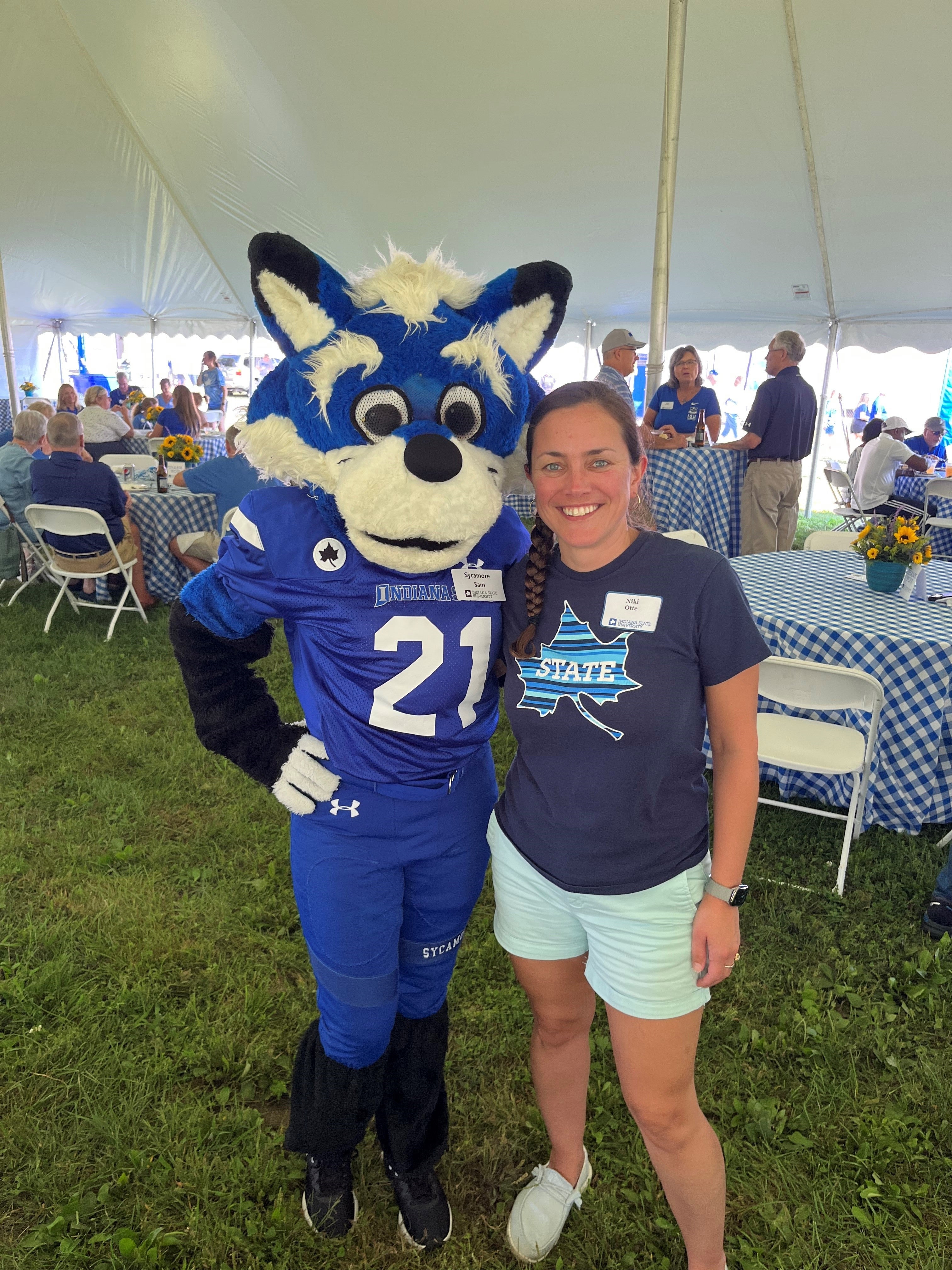 A White woman wearing a navy blue “Sycamores” t-shirt, light blue shorts, and white sneakers poses smiling with a costumed mascot in a blue football uniform under a large white event tent. Tables with blue and white checkered cloths and flower centerpieces are visible in the background.