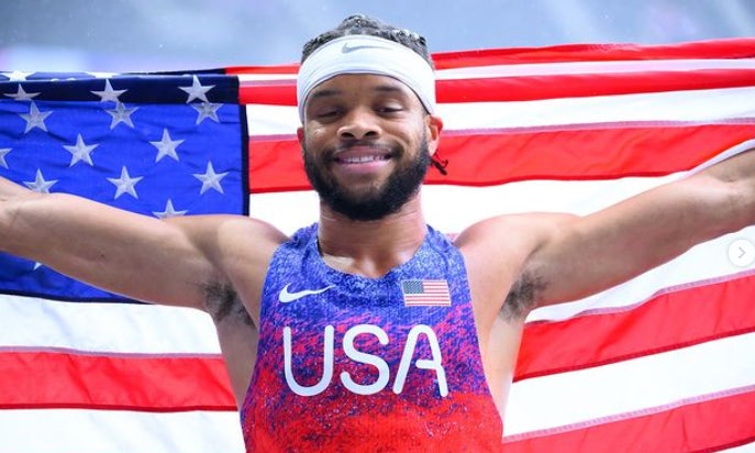 A young Black man stands joyfully holding an American flag outstretched behind him. He wears a white headband and a sleeveless navy athletic uniform with bold “USA” letters across his chest, along with a small American flag patch and a Nike logo.