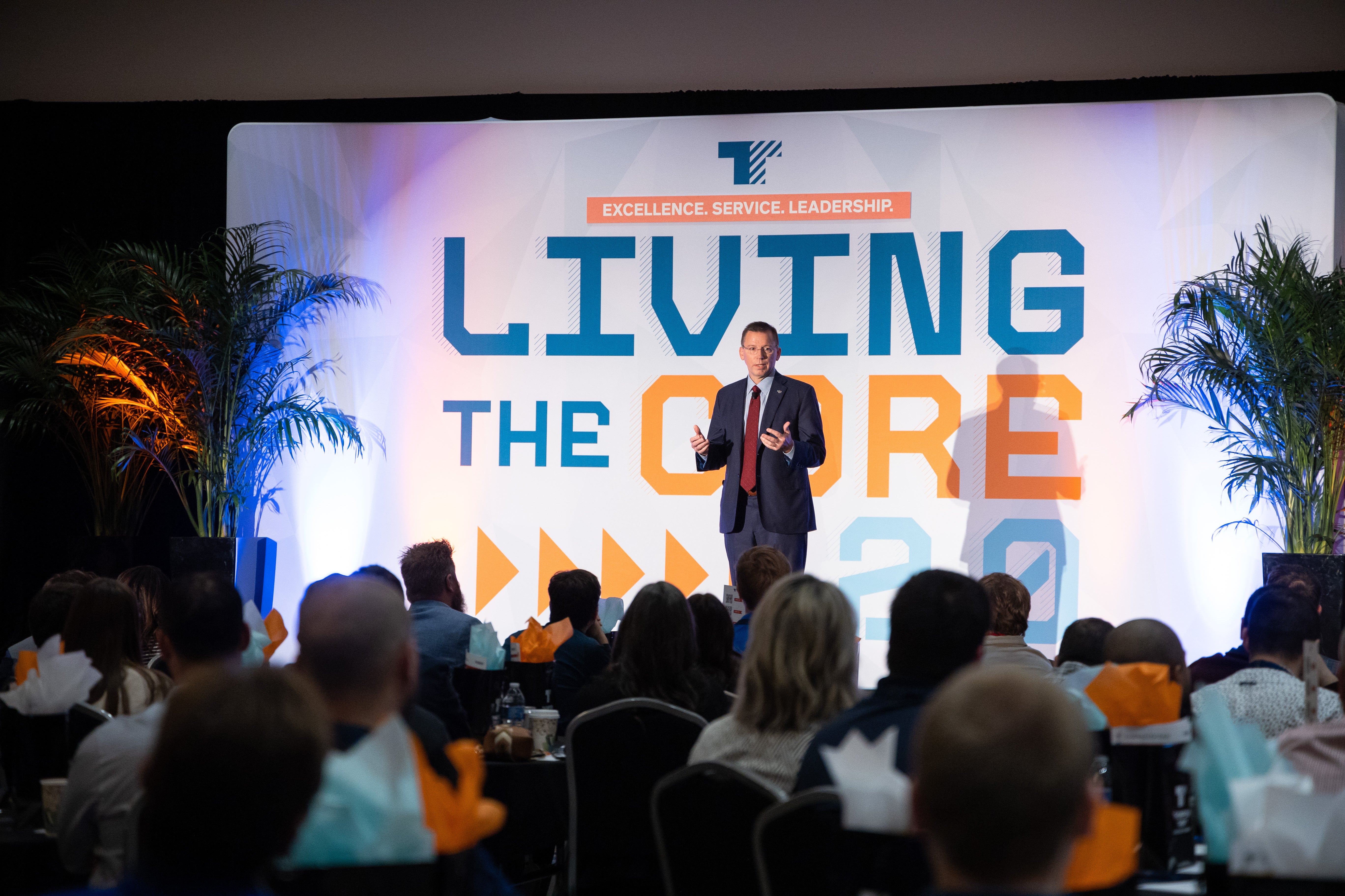 A White man in a dark suit and red tie stands speaking on stage before an audience, with green potted plants on both sides of a backdrop that reads “Living the Core” and “Excellence. Service. Leadership.”