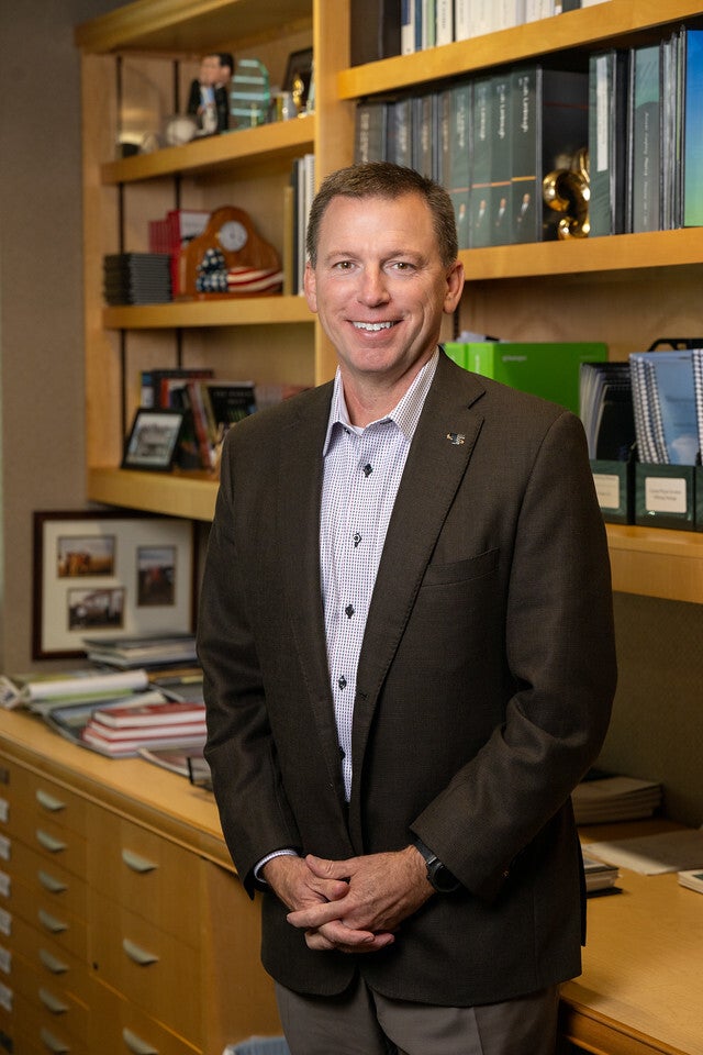 A White man with short brown hair wearing a brown blazer over a light checkered shirt stands smiling in an office lined with shelves of books, framed photos, and decorative items.