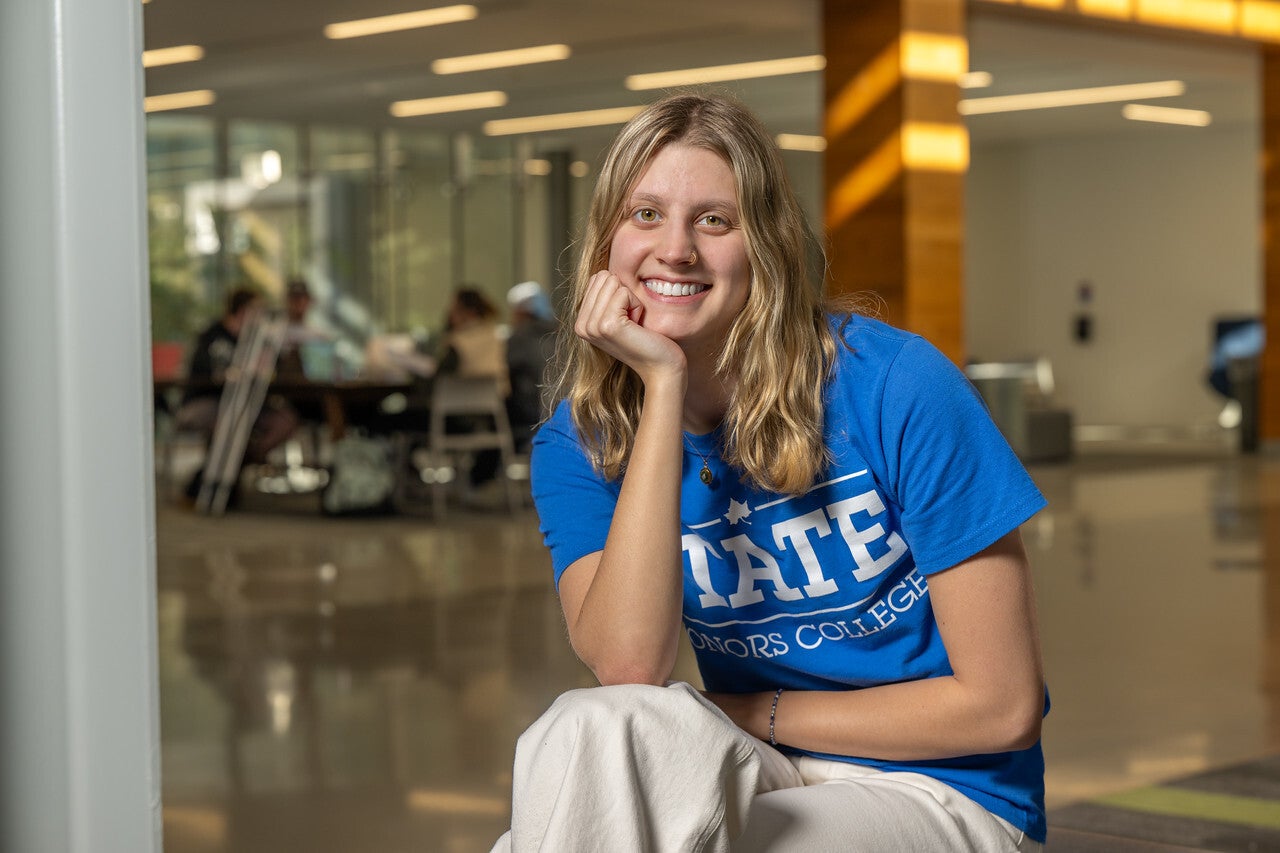 A young White woman with shoulder-length blonde hair smiles while seated indoors, resting her chin on her hand. She wears a blue “STATE Honors College” T-shirt and light-colored pants, with students working at tables in the blurred background.
