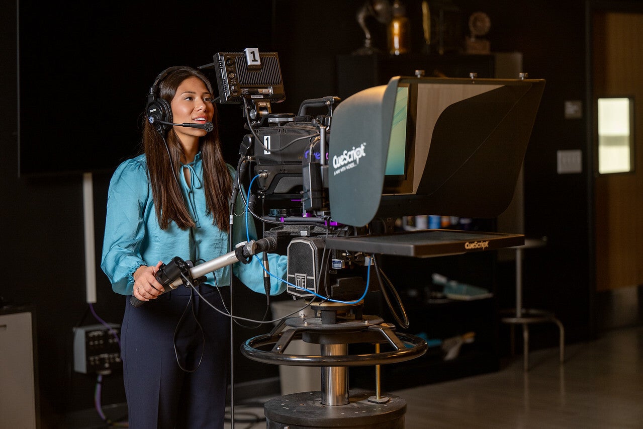 A young woman with long brown hair operates a professional video camera in a studio. She wears a headset, a light blue blouse, and dark pants. The dimly lit background features studio equipment and shelves.