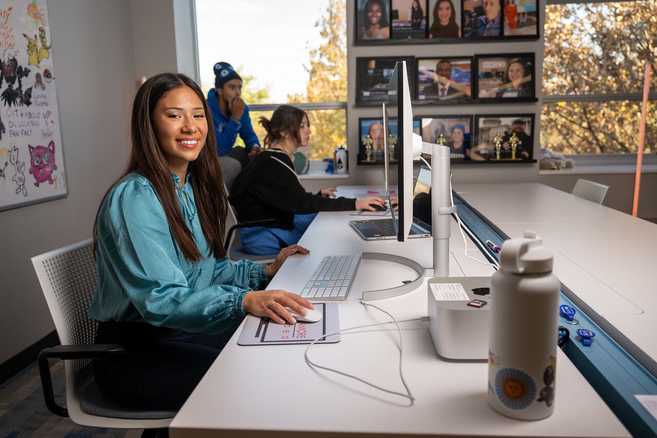 A young woman with long brown hair wearing a light blue blouse sits at a white desk, holding a computer mouse in front of a large monitor. Other people are seen working in the background, and the room features a whiteboard with drawings and framed photos on the wall.