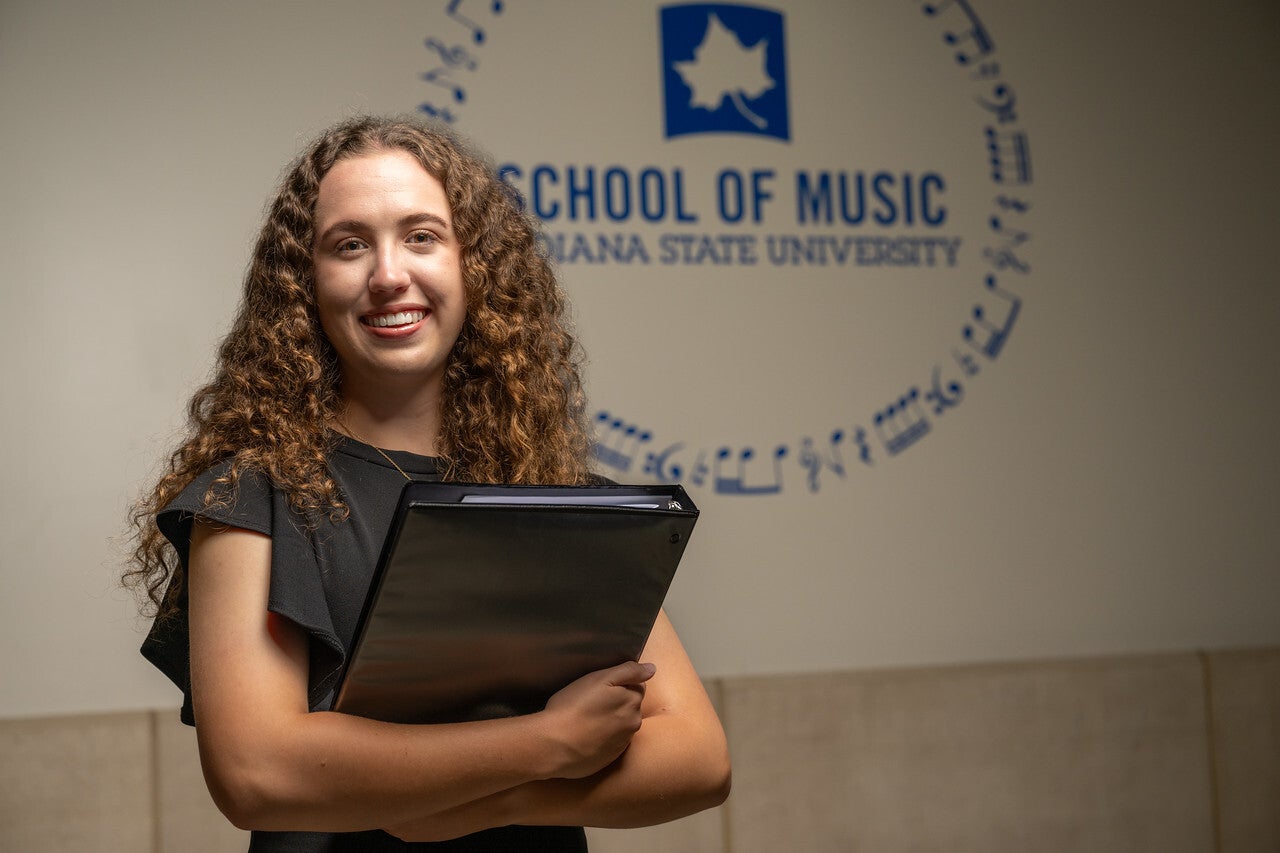 A white woman with long curly hair stands in front of a wall displaying the logo and text for the School of Music at Indiana State University. She is smiling and holding a black binder, dressed in a black outfit.