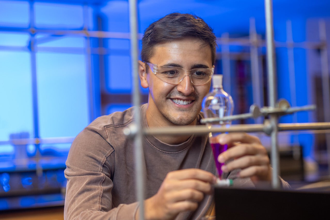A young man wearing safety glasses and a brown long-sleeve shirt smiles while working with a glass flask containing a purple liquid in a laboratory with blue lighting and metal lab equipment.