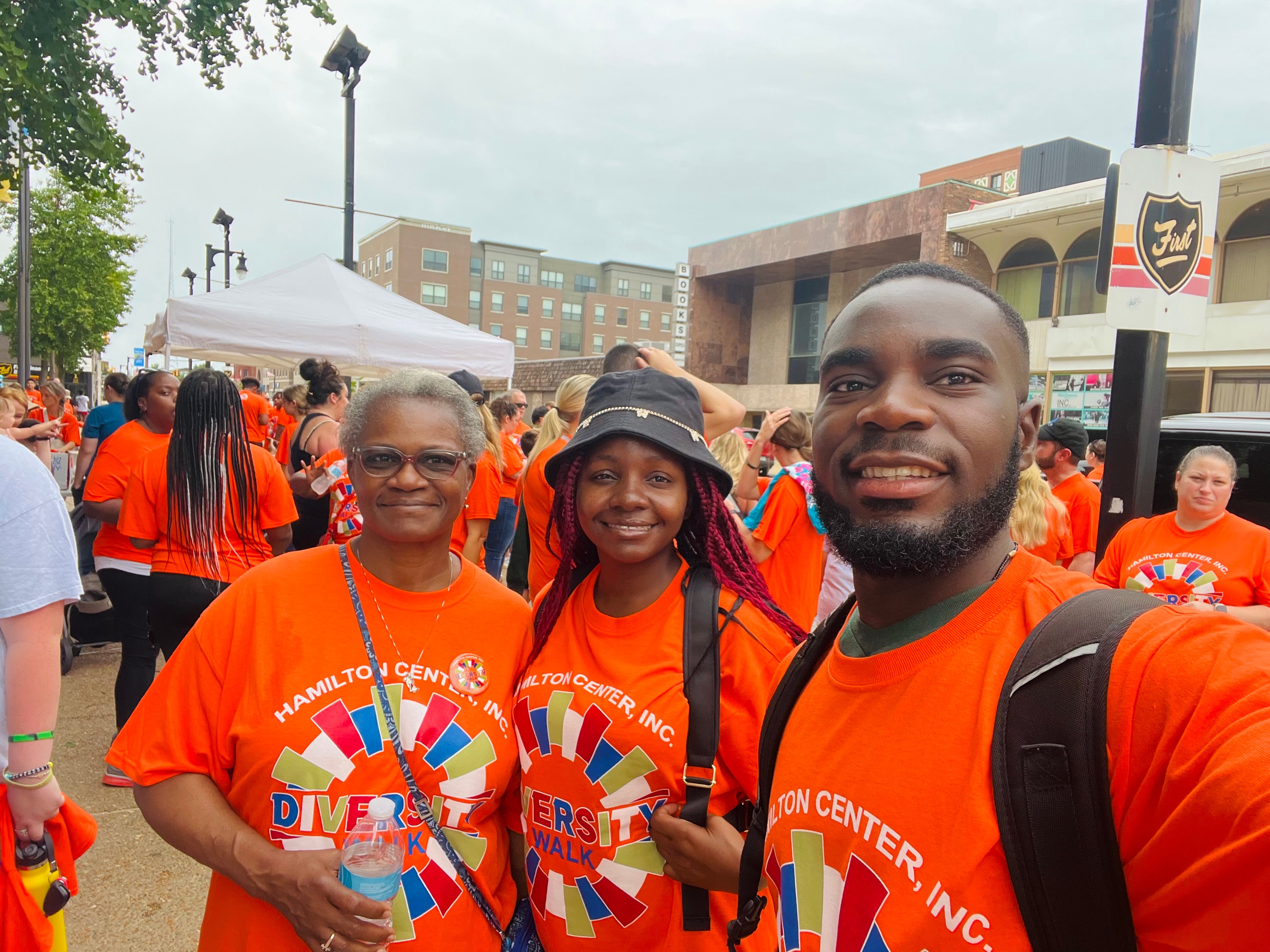Three smiling Black individuals stand together at an outdoor event wearing bright orange “Hamilton Center, Inc. Diversity Walk” T-shirts. The older woman on the left holds a water bottle, the young woman in the middle wears a black bucket hat, and the man on the right carries a backpack.