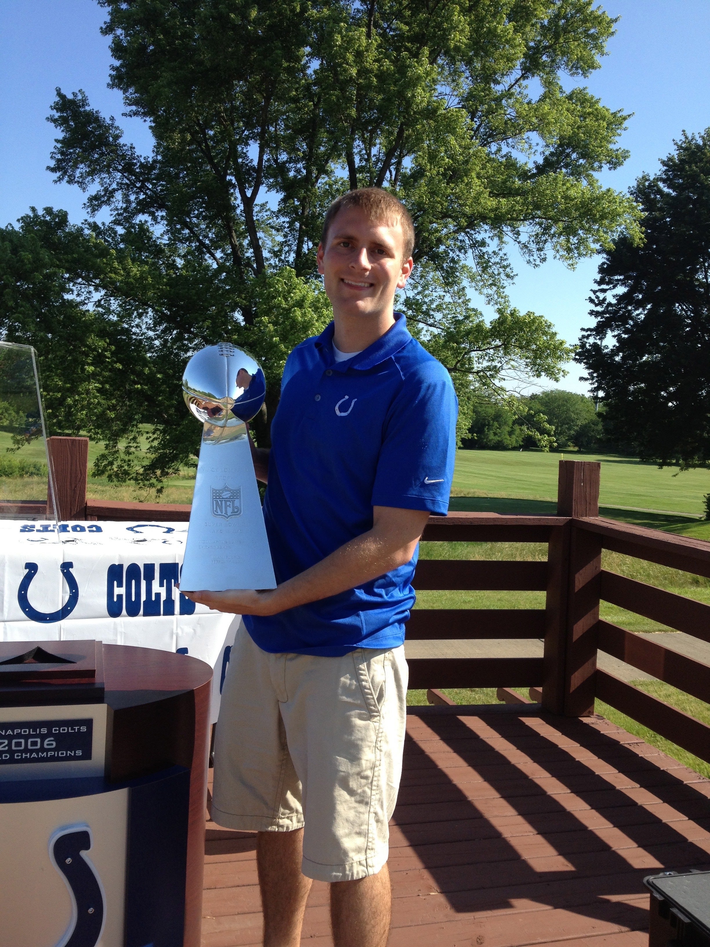 A smiling man in a blue Indianapolis Colts shirt and khaki shorts holds a large silver NFL trophy outdoors. He stands on a wooden deck with a Colts display table behind him, surrounded by green trees and a clear blue sky.