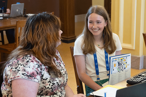 wo young women are seated at a table in a library or study area, smiling at each other as they review a document with graphics and text. The woman on the left, with straight, shoulder-length, blondish-brown hair, holds the document, which appears to have a colorful map on the visible side. The other woman has curly, dark-brown, shoulder-length hair. The table has computers and books, indicating a collaborative academic or work setting.
