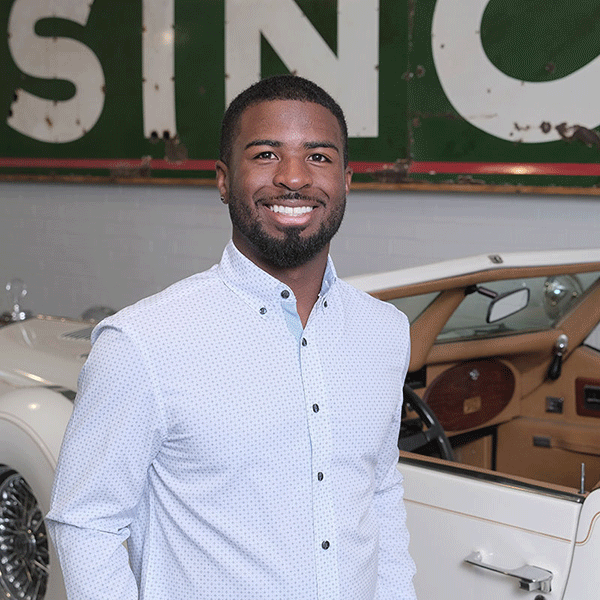 A Black man wearing a light button-down shirt stands smiling indoors in front of a white vintage car, with a large, weathered sign hanging on the wall behind him.