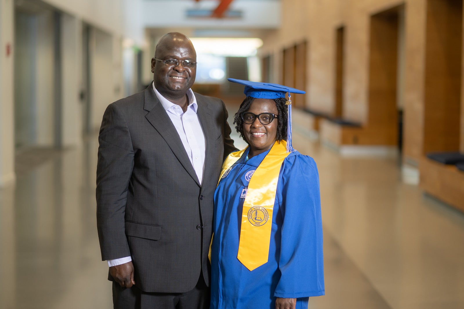 A Black man in a dark suit and white shirt stands beside a Black woman wearing a blue graduation gown, cap, and yellow stole. Both are smiling and standing in a bright hallway with soft light and blurred background.