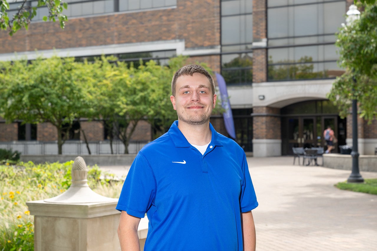 A white man with short light brown hair and a trimmed beard stands outdoors wearing a bright blue collared shirt. Behind him are green trees, a tan stone railing, and a brick building with large windows.
