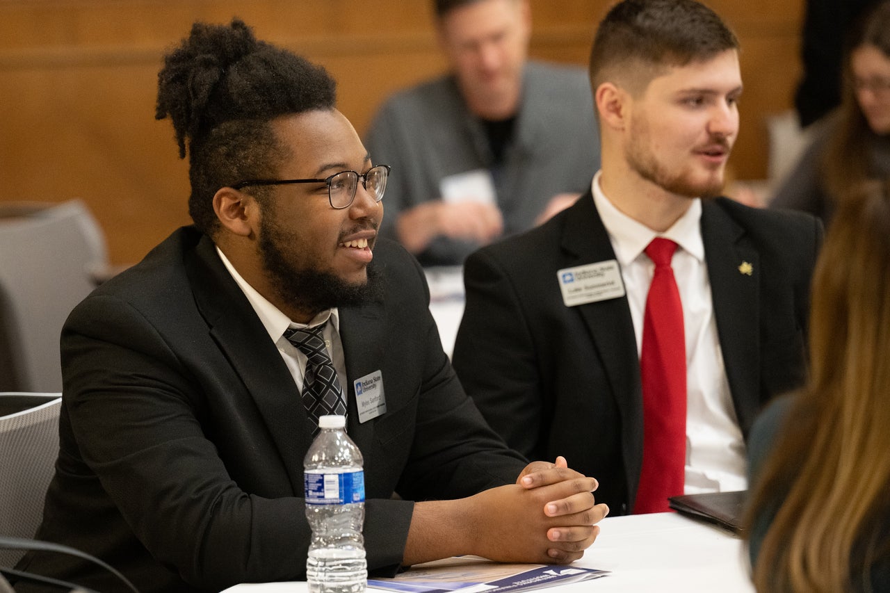 A Black man with short twists, glasses, and a beard sits at a table wearing a dark suit and patterned tie, smiling. Beside him, a white man with short brown hair and a trimmed beard wears a suit with a red tie. Other people are visible in the background.
