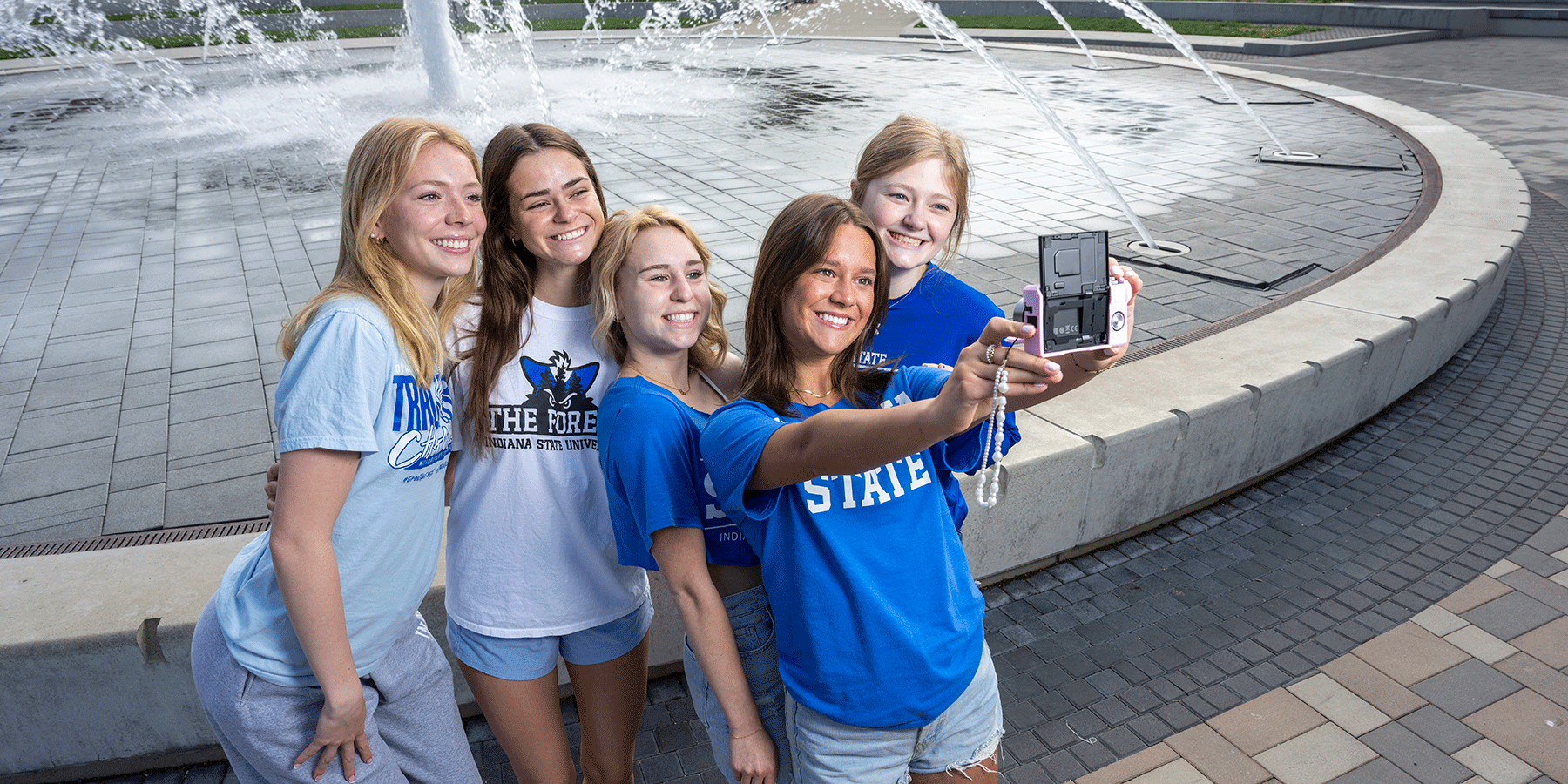 Five smiling female students taking a selfie in front of a large fountain with water jets. They wear Indiana State shirts and casual summer clothing.