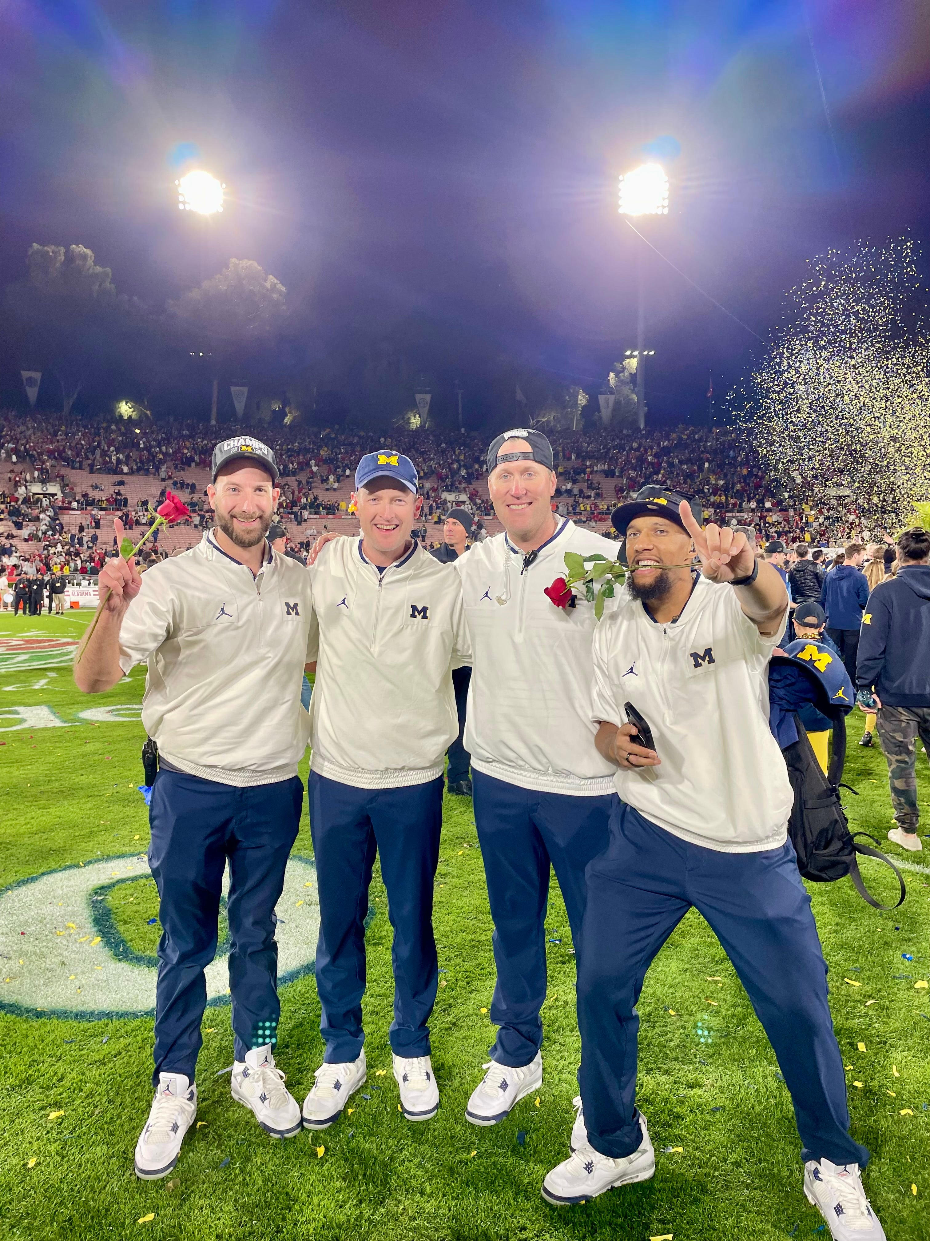 Four men stand smiling on a football field at night, dressed in matching white Michigan team jackets, navy pants, and white sneakers. Two hold red roses, and one flashes a number-one gesture as confetti falls behind them. Stadium lights and a cheering crowd are visible in the background.