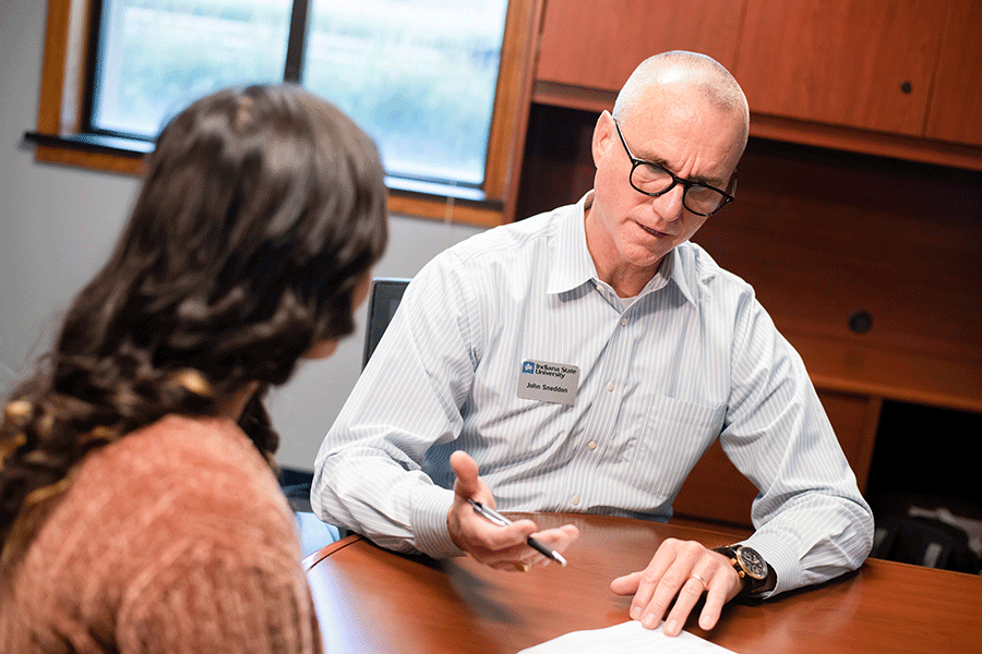 Two people sitting at a wooden desk in an office setting. One, wearing a striped button-up shirt, gestures toward papers while holding a pen. The other person listens attentively. A wooden cabinet and a sunlit window are in the background.