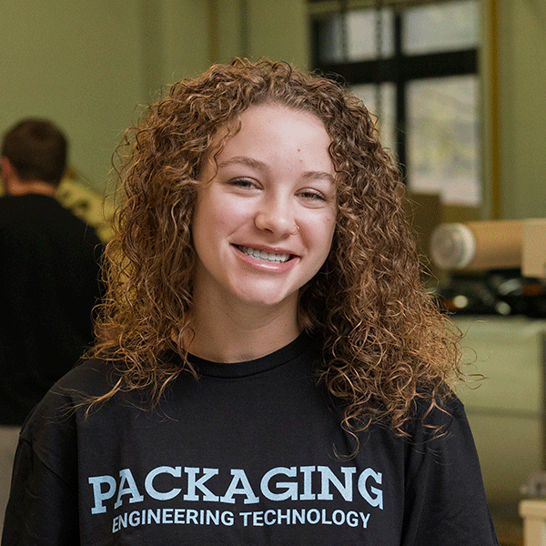 A young white woman with long, curly light brown hair, wearing a black T-shirt that reads “Packaging Engineering Technology” in light blue text, smiles while standing in a room. The background is softly blurred, with another person and equipment visible behind her.