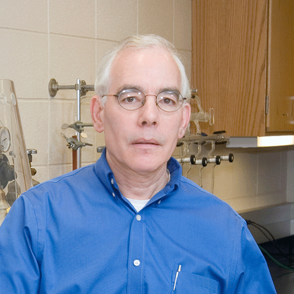 An older white man with short white hair and round glasses, wearing a blue button-down shirt, stands in a laboratory setting. Behind him are lab fixtures, glass equipment, and wooden cabinets.