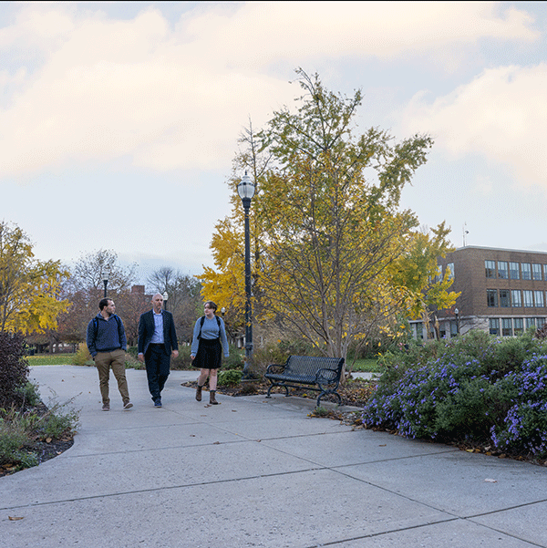 Three people walk along a paved pathway in an outdoor campus setting with autumn foliage. Trees with yellow leaves, bushes, and flowers line the path. A bench and a lamppost are visible, with a multi-story building featuring large windows in the background.