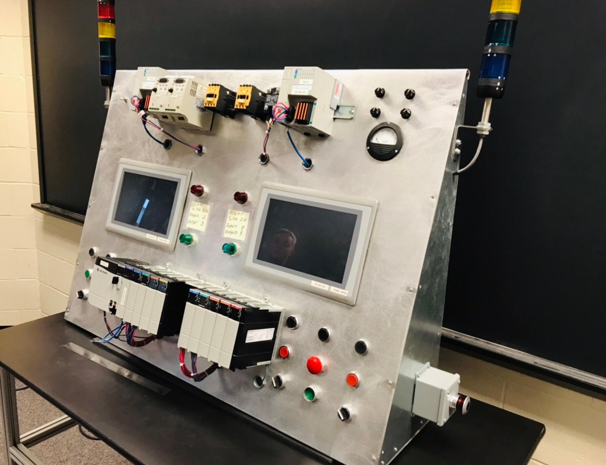 A large industrial control panel sits on a table in a classroom, featuring dual touchscreens, buttons, indicator lights, wiring components, and mounted programmable logic controllers. Two vertical signal tower lights are attached to the top corners of the metal panel, and a black chalkboard is in the background.