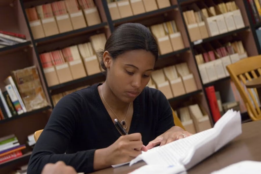 A Black female student wearing a black long-sleeved shirt sits at a wooden table in a library or study room, writing in a notebook. Shelves filled with books and files are visible in the background, and an empty wooden chair is nearby.