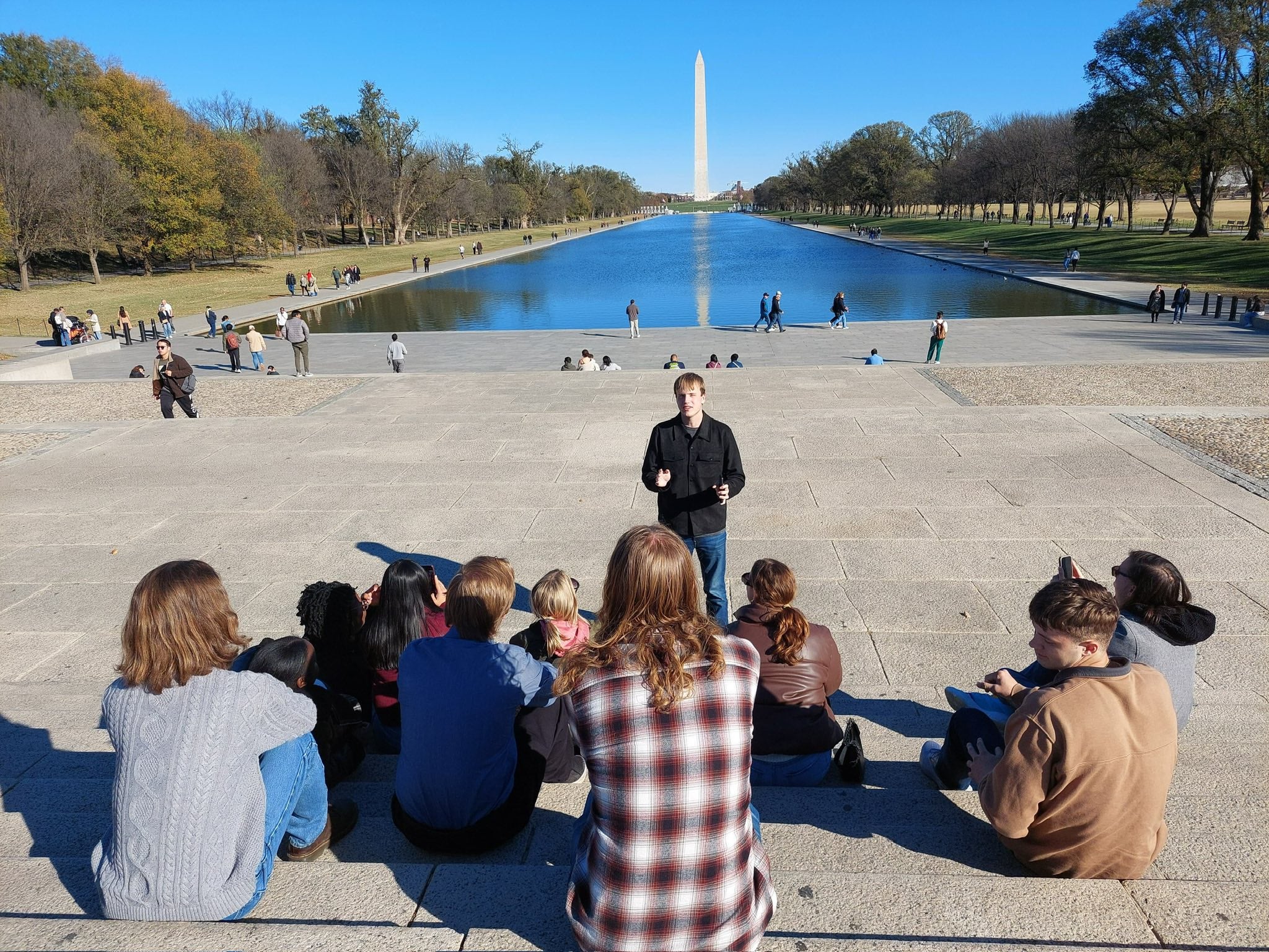 A group of people sits on the steps overlooking the Reflecting Pool in Washington, D.C., listening to someone standing and speaking in front of them. The Washington Monument rises in the distance under a clear blue sky, and visitors walk along the pool and nearby paths.