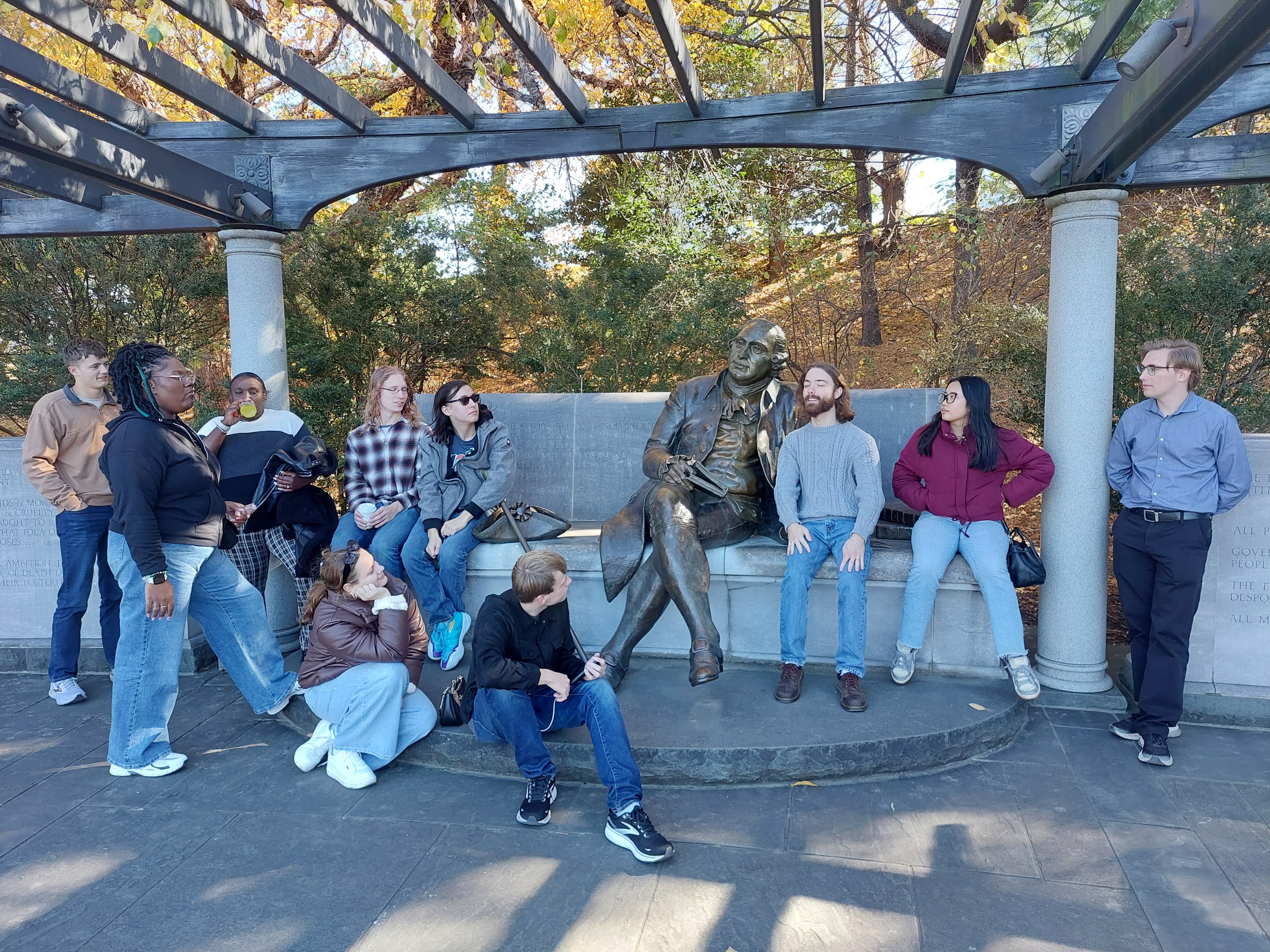 A group of people gathers around a bronze statue of President Franklin D. Roosevelt seated on a bench at the FDR Memorial in Washington, D.C. Several members of the group sit or lean on the bench beside the statue, while others stand nearby under a wooden pergola.
