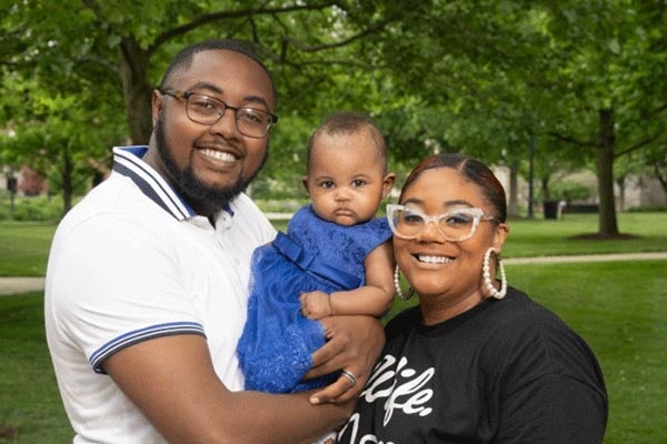 A family stands together outdoors in a green park, with two adults holding a baby between them. Trees and grass in the background create a calm, natural setting, and the group appears relaxed and close.