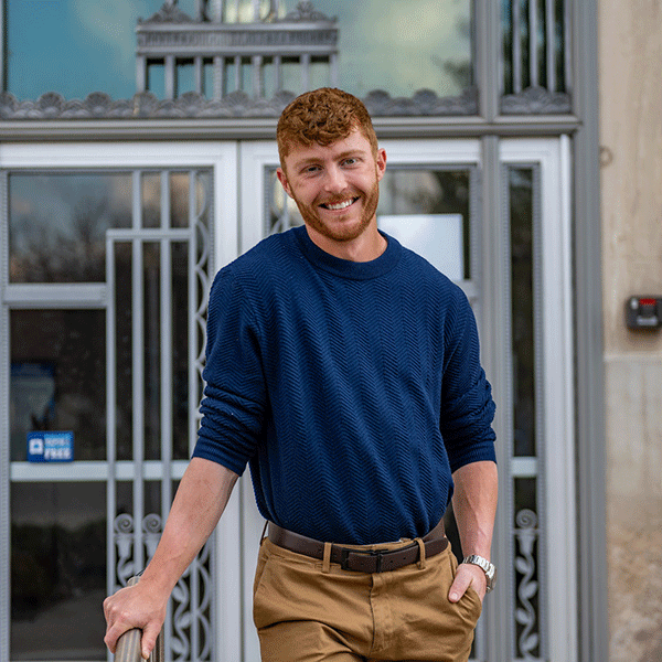 “Smiling man with curly hair and a beard wearing a blue sweater stands outside in front of a building entrance.”
