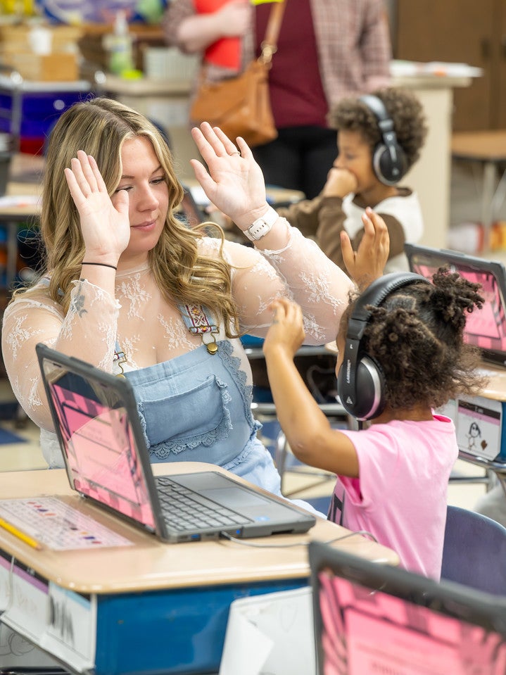 A white female with blonde hair sitting at a student desk raises her hands in a playful or celebratory gesture while interacting with a young child wearing headphones. Laptops with pink screens are open on the desks, and other children work in the classroom in the background.