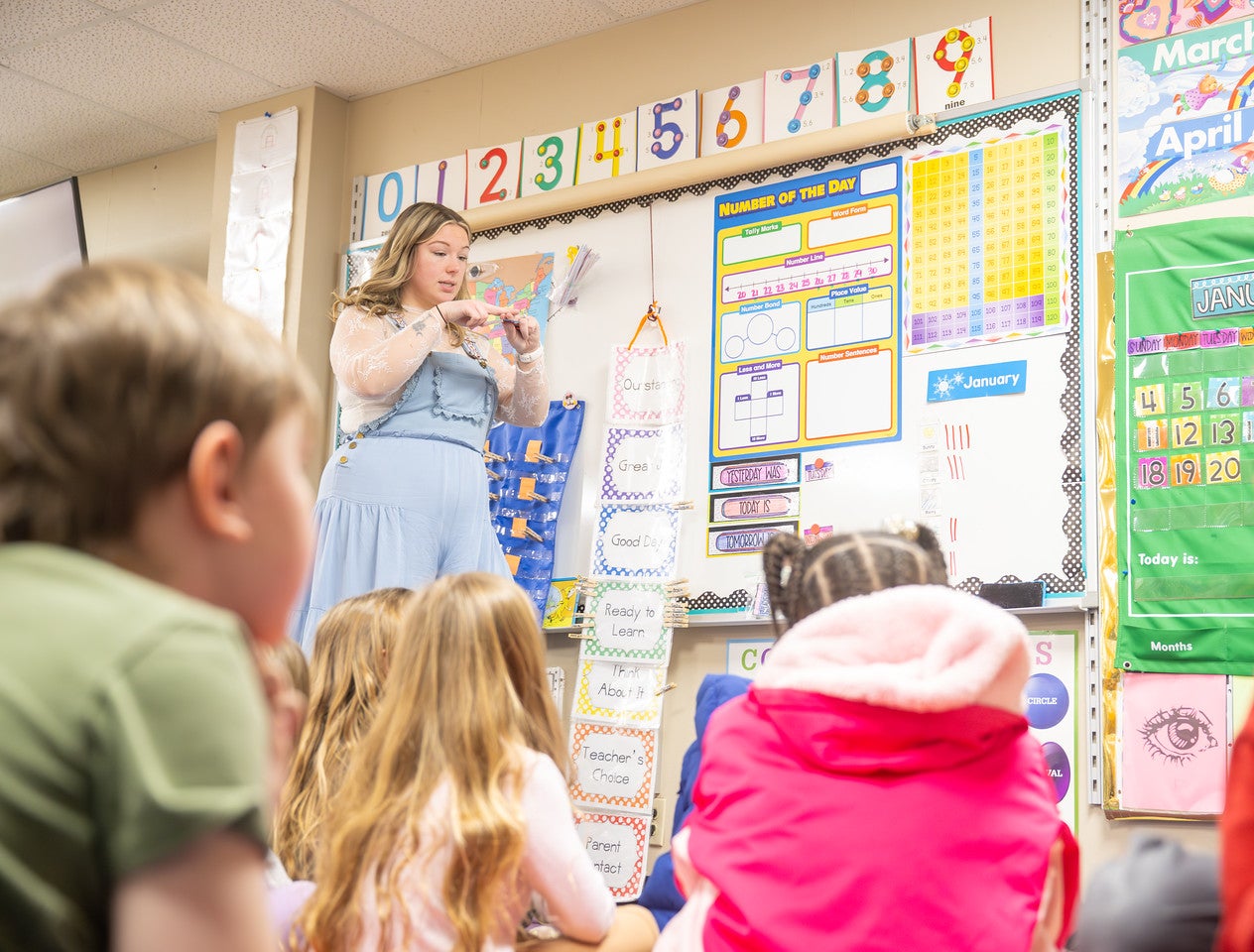 A white female teacher with blonde hair stands at the front of a brightly decorated early‑elementary classroom, holding a small object while a group of young students sit on the floor watching. Colorful number charts, calendar pieces, and educational posters fill the wall behind her. The teacher wears light blue overalls over a white sheer lace top. 