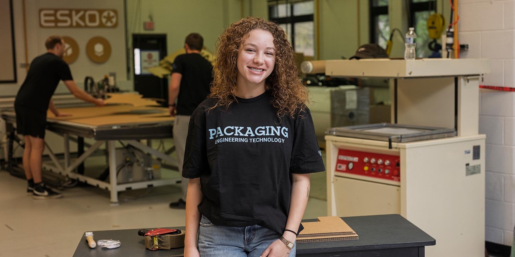 Student wearing a black “Packaging Engineering Technology” T-shirt smiles while standing in a packaging lab, with cardboard sheets, industrial equipment, and two people working at tables in the background.