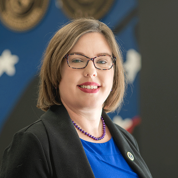 “Woman wearing glasses, a blue top, and a black blazer smiles in an indoor portrait with a blurred background.”