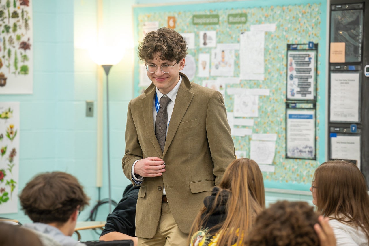 A classroom scene with a white male with curly brown hair wearing a brown blazer and tie stands near student desks, engaging with a group of students. Educational posters, papers, and a bulletin board line the walls.