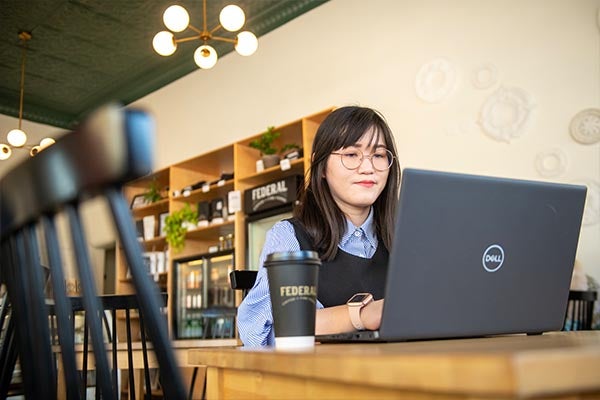A young woman with shoulder-length black hair sits at a wooden table in a café-style space, working on a laptop with a coffee cup beside it. Shelving, plants, and warm lighting in the background create a comfortable, informal study environment.